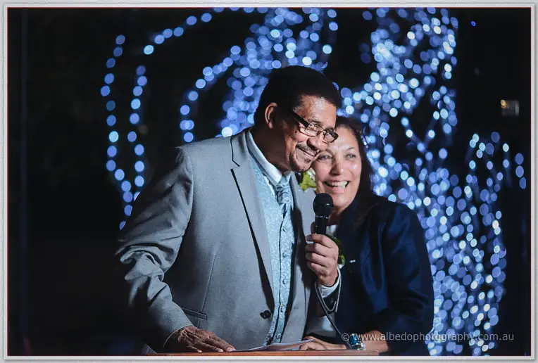 A guest couple leans into a single microphone, laughing beneath the glittering blue canopy.