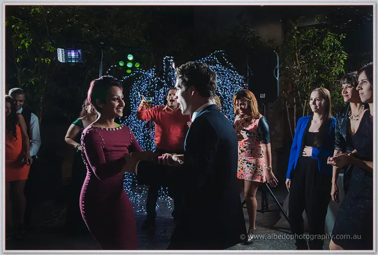 Guests dance under party lights, a woman in maroon twirling with a man in a dark suit among fairy-lit trees