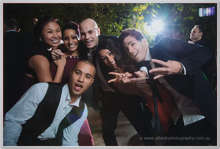 Wedding guests crowd together for a playful group pose, flashing peace signs under the courtyard lights