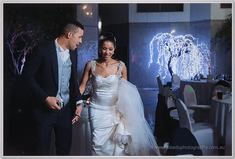 Cristal and Troy stroll hand in hand past their reception tables beneath the glowing fairy-lit willow
