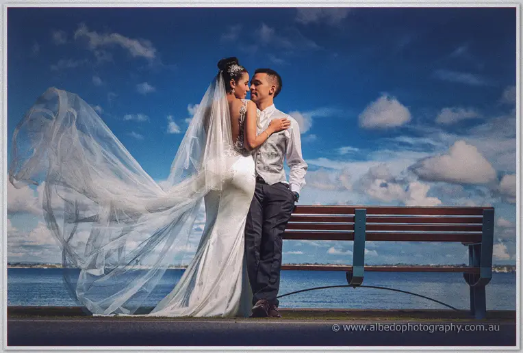 Cristal and Troy lean on a bench by the water beneath a dramatic blue sky as her long veil sweeps in the wind