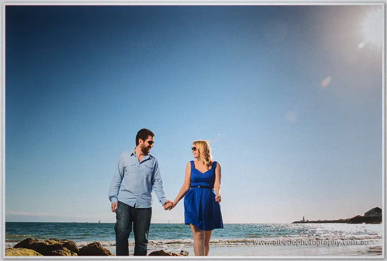 Julie and Dale walk hand in hand along the shoreline in matching blue, sun high over the bay