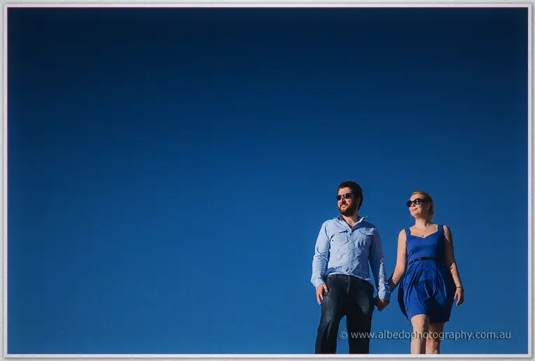 In sunglasses, the couple stroll barefoot along the sand beneath a vast cobalt sky
