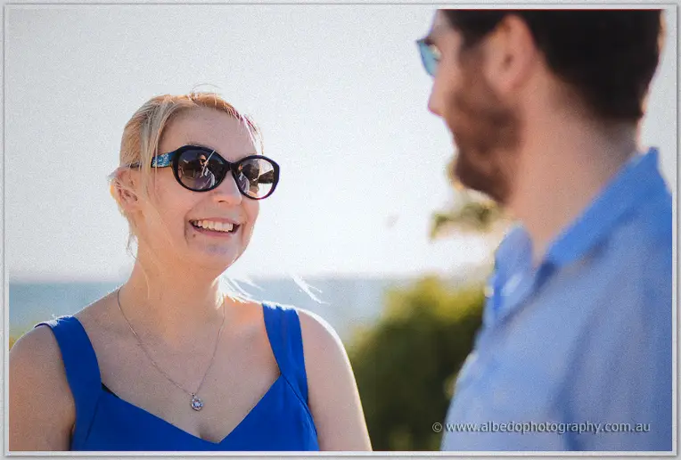 Julie laughs warmly over her sunglasses as Dale, softly blurred, faces her by the sea