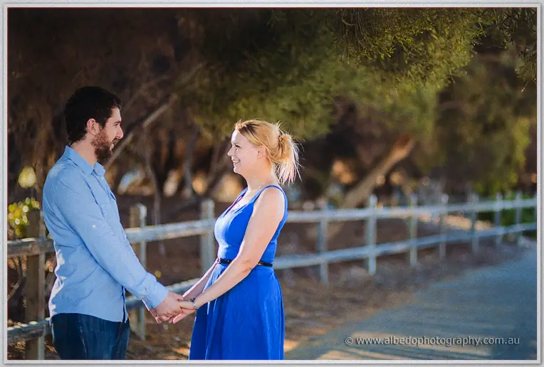 Holding both hands on a quiet path, the couple smile at each other beside a dappled fence