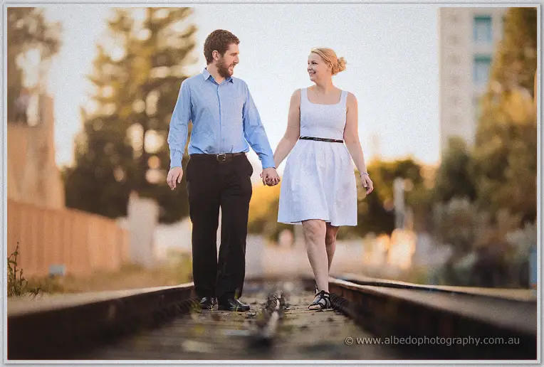 Julie in a white dress and Dale stroll hand in hand down overgrown railway tracks at sunset