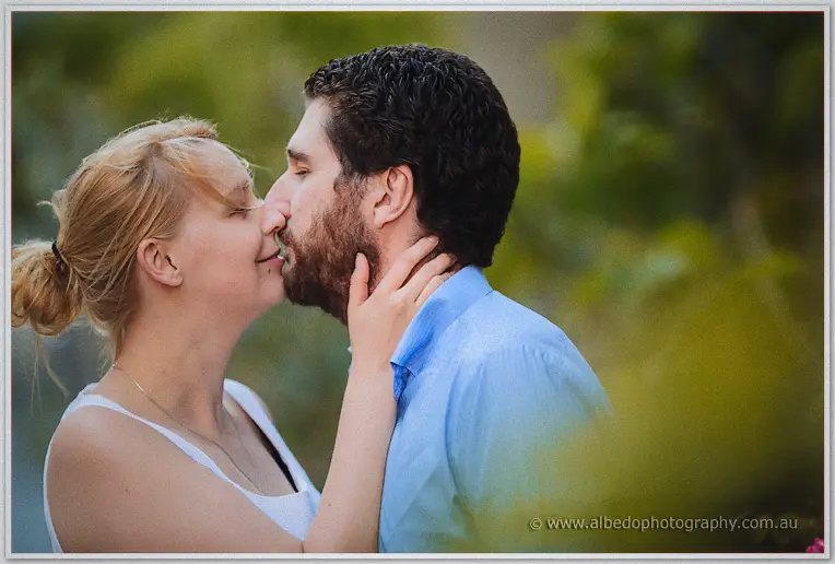 Julie touches Dale's beard as they share a tender kiss against soft green foliage