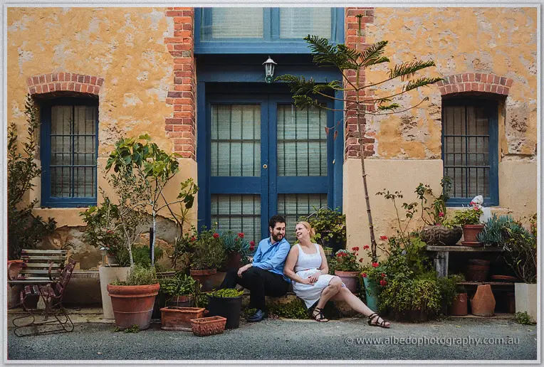 Julie and Dale sit side by side among potted plants before a blue door on a ochre wall