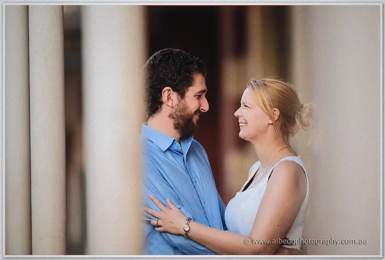Seen between pale stone columns, Julie rests a hand on Dale's chest as they beam at each other