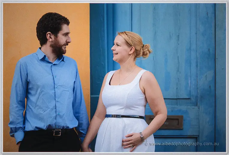 Smiling at each other beside a weathered blue door, warm ochre wall at their backs