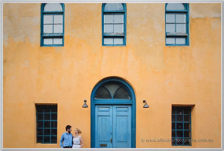 Dwarfed by a sun-faded ochre facade with teal windows, the couple chat quietly below
