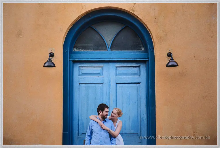 Julie wraps her arms around Dale under a grand arched blue doorway on an ochre wall