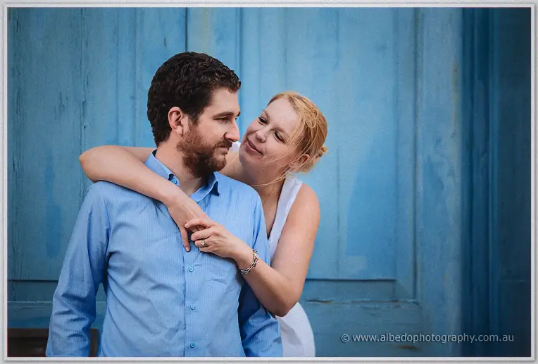Leaning over Dale's shoulder in front of a weathered blue timber door, Julie smiles down at him