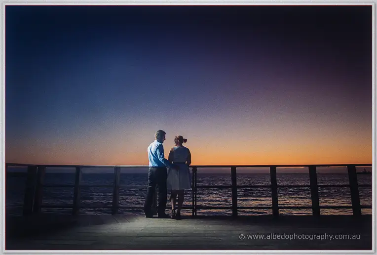 Julie and Dale stand close at the jetty rail as dusk fades from amber to deep blue over the sea