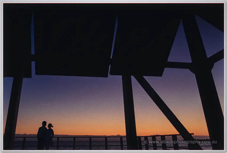 A small silhouetted couple lean on the jetty rail under dark beams at deep dusk