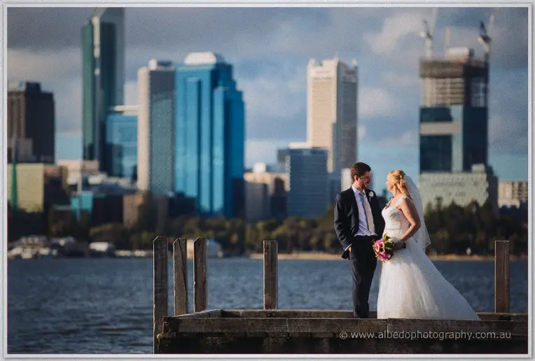 Julie and Dale pause on a river jetty, the Perth towers soft across the water behind them