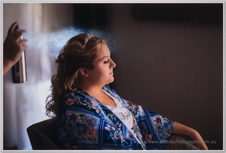 Hairspray mist drifts around Olivia's curls and tiara in her floral robe as final touches settle in.