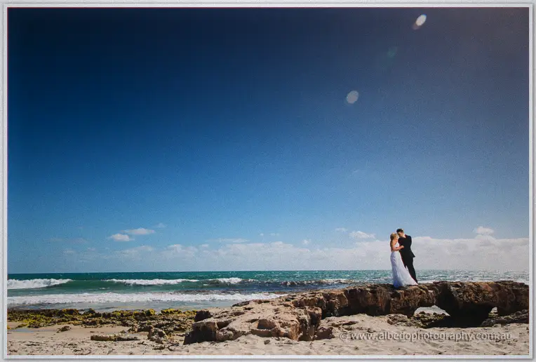 Olivia and Max embrace on a rocky outcrop beneath a vast blue sky, waves rolling behind them.