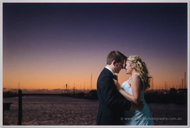 Olivia and Max press foreheads together at the marina, silhouetted against a violet and orange dusk.