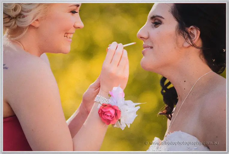 A bridesmaid touches up Sarah's lipstick outdoors, both laughing against sunlit golden foliage.