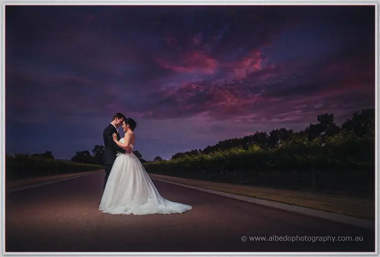Sarah and Chris embrace on a tree-lined drive as purple and magenta clouds glow at dusk.