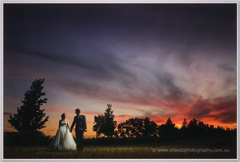 Sarah and Chris stand hand in hand on a lawn at Sandalford under a blazing pink and orange sunset.