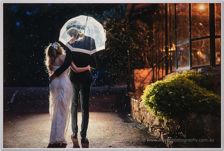 Tara and Stephen share an umbrella under the falling rain past the softly lit stone walls