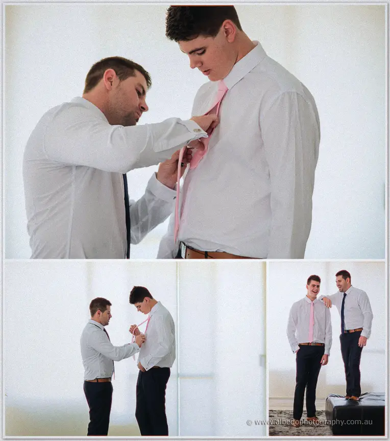 A groomsman threads and knots a pink tie for a friend in a bright, sunlit white room.