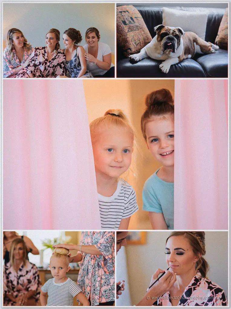 Two little girls peek through pink curtains grinning as makeup and hair touch-ups finish nearby.