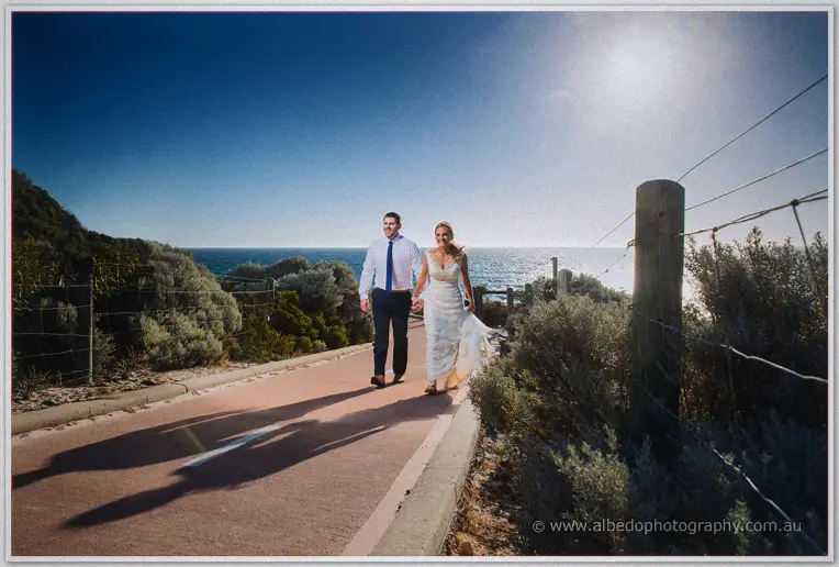 Tarmecca and Lee stroll a coastal path above the ocean, the afternoon sun throwing long shadows