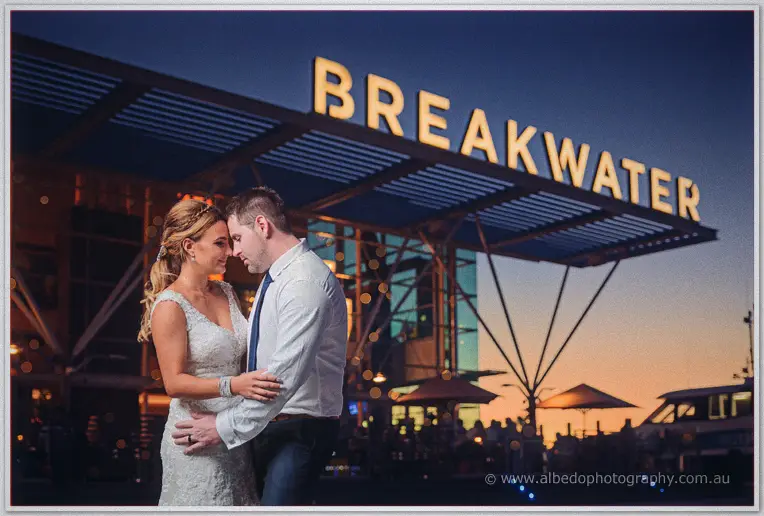 Tarmecca and Lee embrace at dusk beneath the glowing Breakwater sign, marina lights sparkling behind