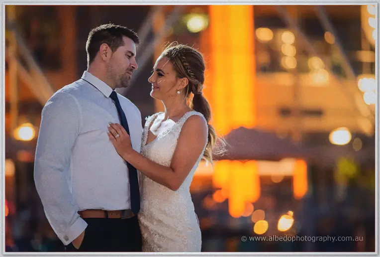 Tarmecca rests her hand on Lee's chest as warm bokeh glows around the evening harbour
