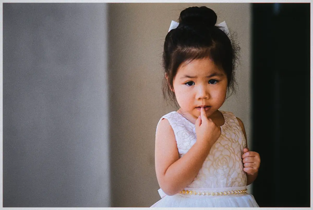 A young flower girl in a white dress looks thoughtfully at the camera with a finger to her lips