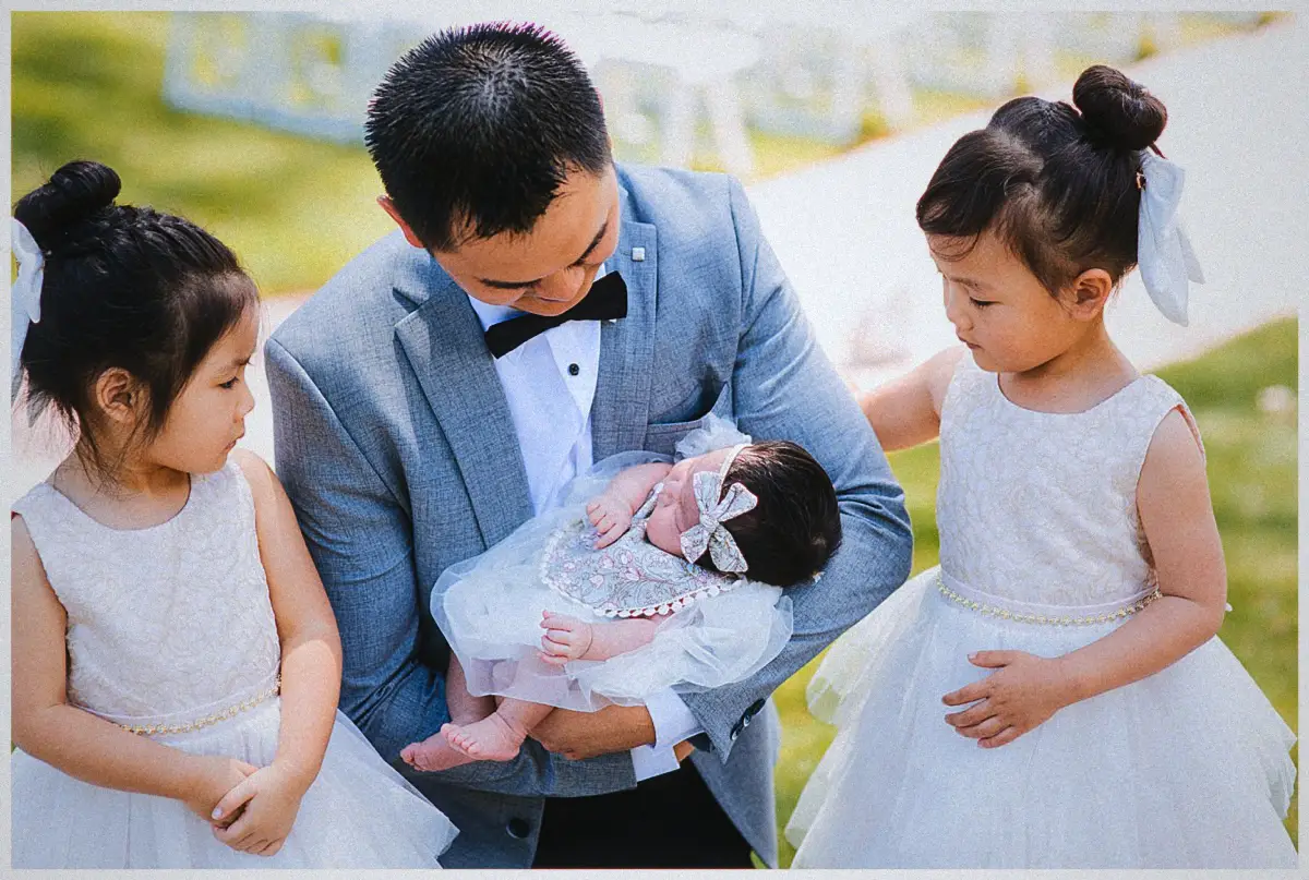 Andrew cradles a newborn on the garden lawn while two flower girls watch closely