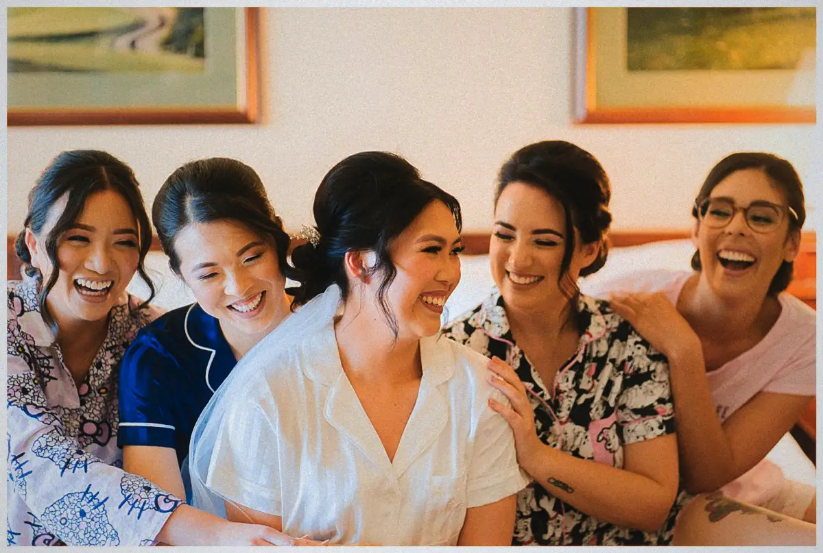 Natalie in her white robe and veil laughs on the bed with her bridesmaids in pyjamas