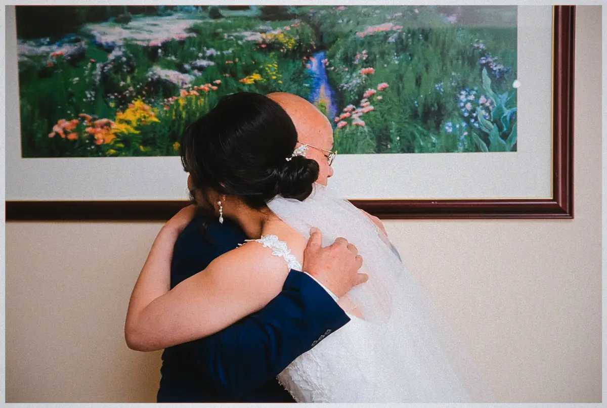 Natalie hugs her father tightly in a navy suit under a framed garden painting