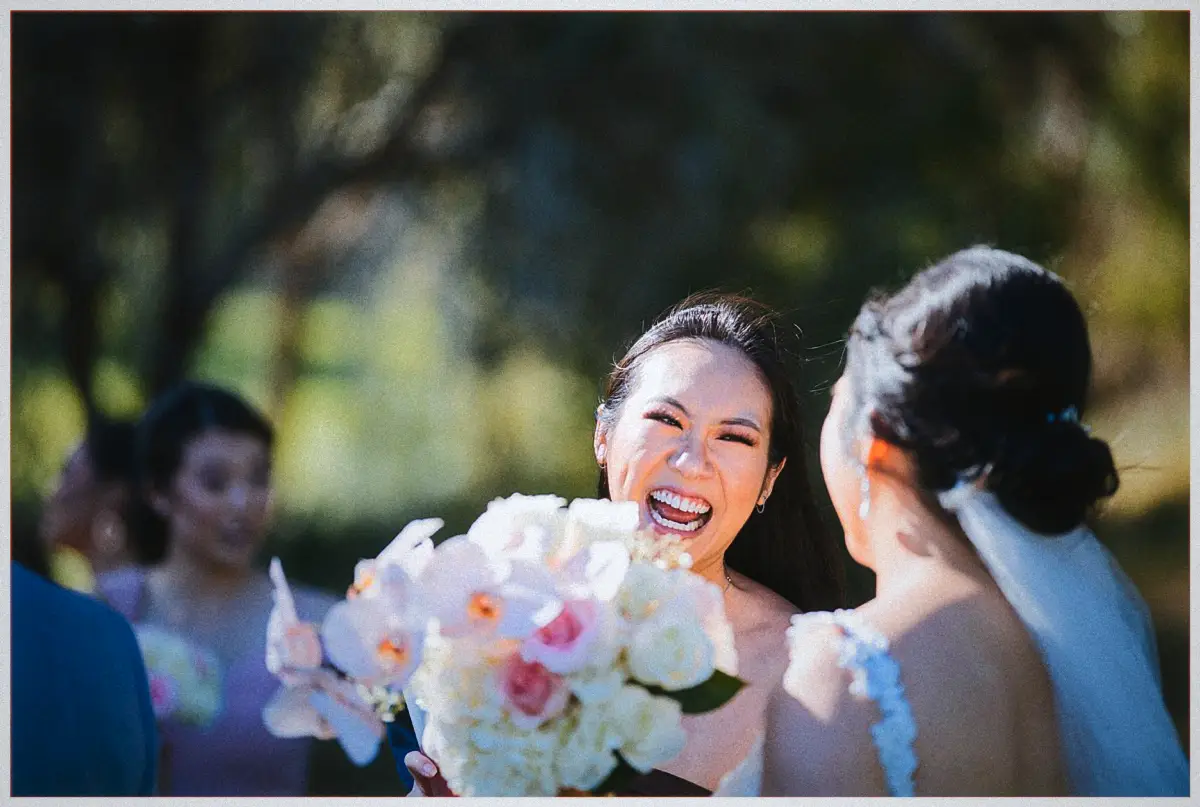 A bridesmaid in blue laughs brightly behind a bouquet of roses and orchids as Natalie turns to greet her