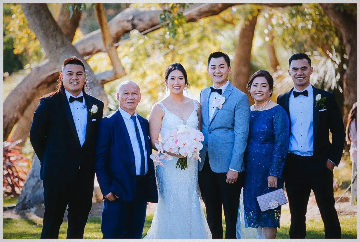 Natalie and Andrew stand with family in suits and a blue lace dress for portraits under the gum trees