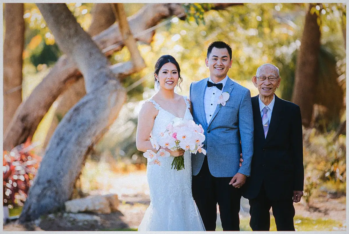Natalie and Andrew flank a smiling older relative in a dark suit for a family portrait in dappled sunlight
