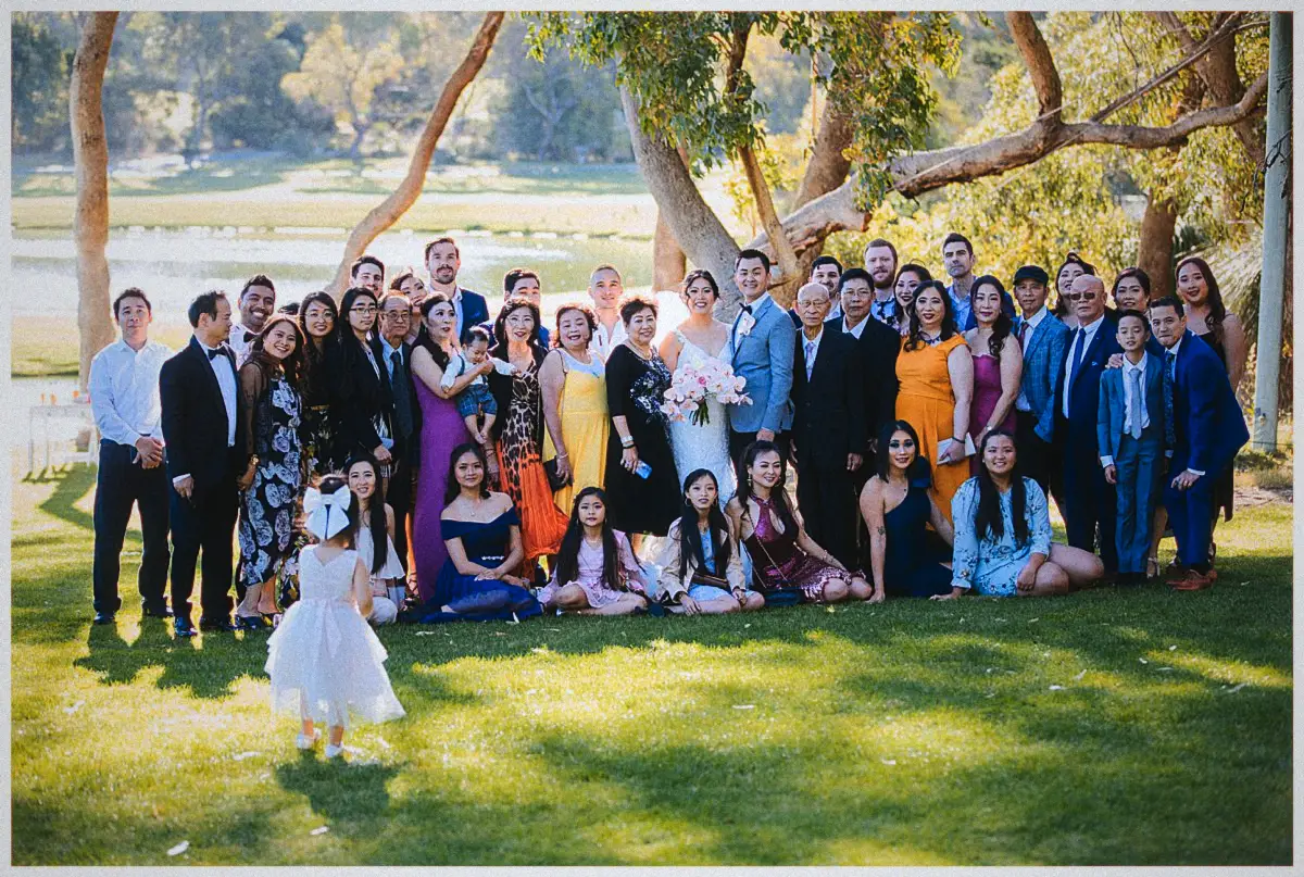 Natalie and Andrew smile at the centre of a large family group as a flower girl wanders onto the lawn