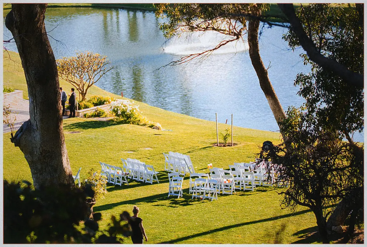 White ceremony chairs wait on the lakeside lawn beneath a spraying fountain at golden hour