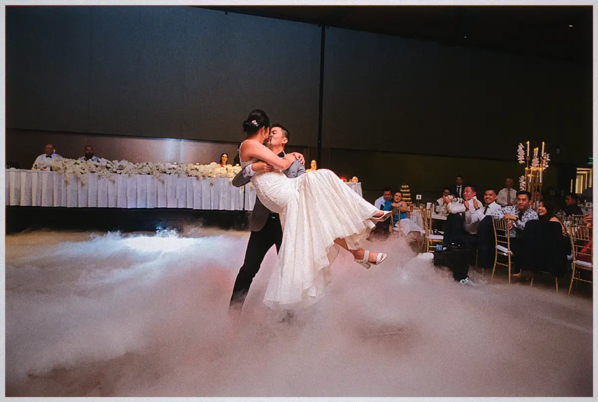 Andrew sweeps Natalie up into a kiss above white fog as guests cheer from candlelit reception tables.
