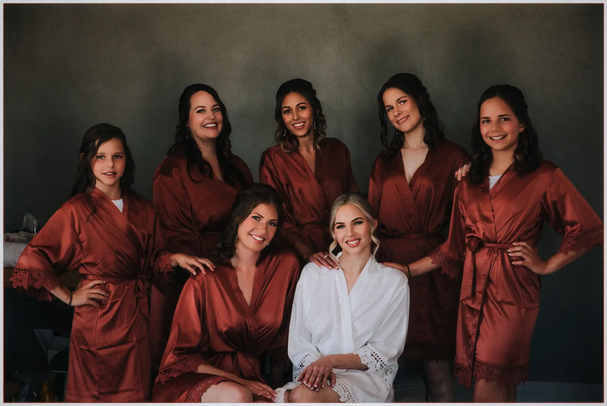 Erin poses with her bridesmaids and junior bridesmaids in rust satin robes against a green wall.