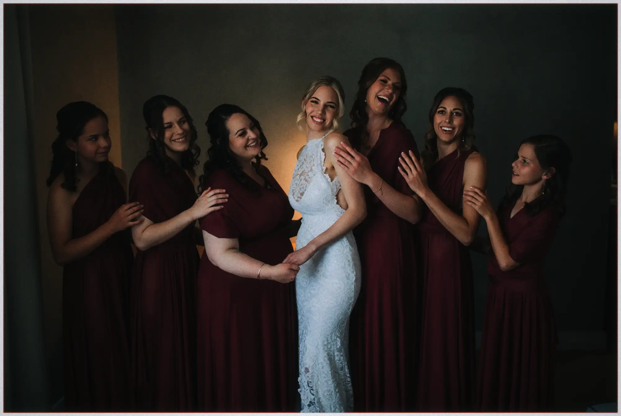 Erin laughs surrounded by her bridesmaids in flowing burgundy gowns, hands resting on her shoulders.