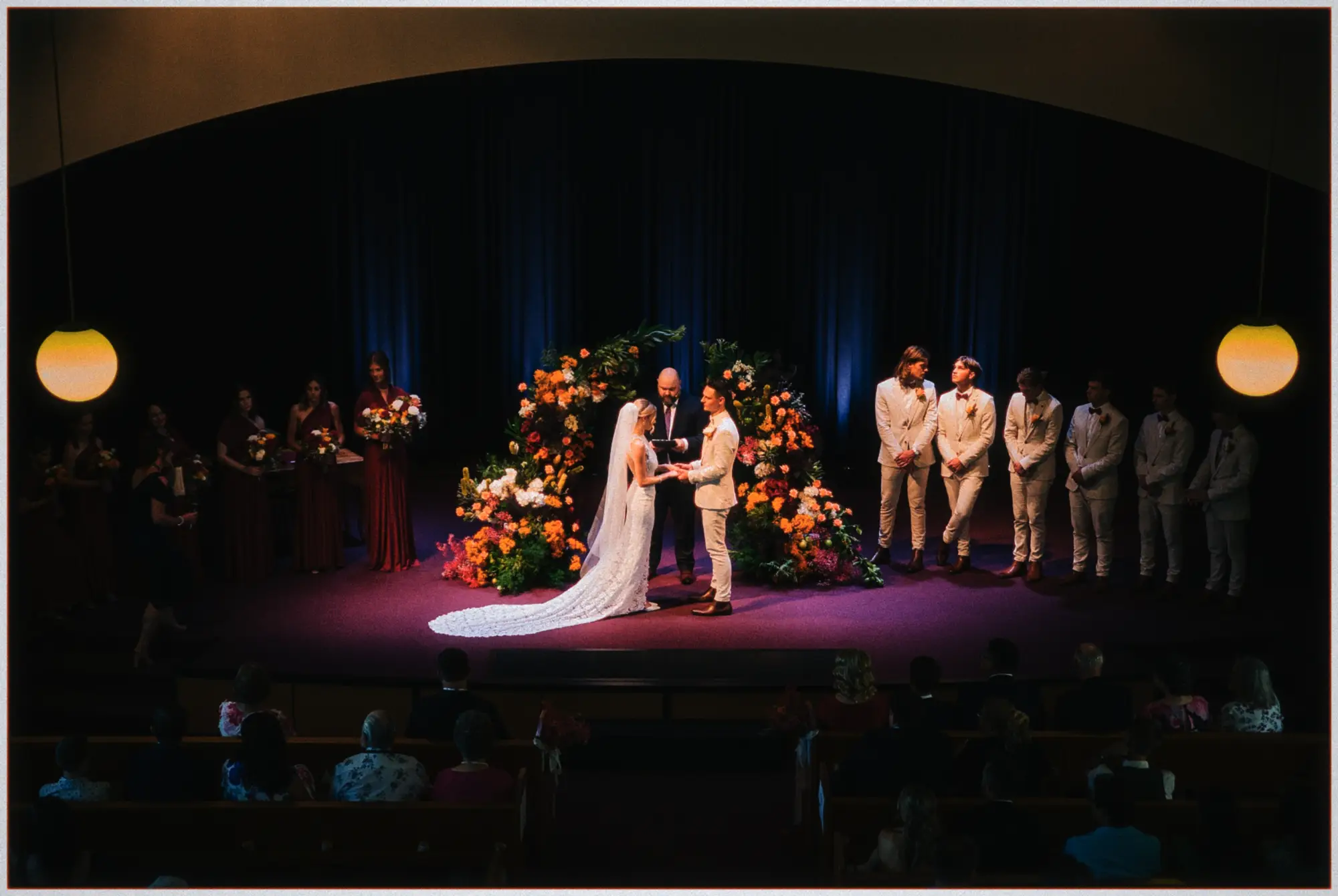 Wide chapel stage view of Erin and Justin marrying between orange blooms and warm hanging lamps.