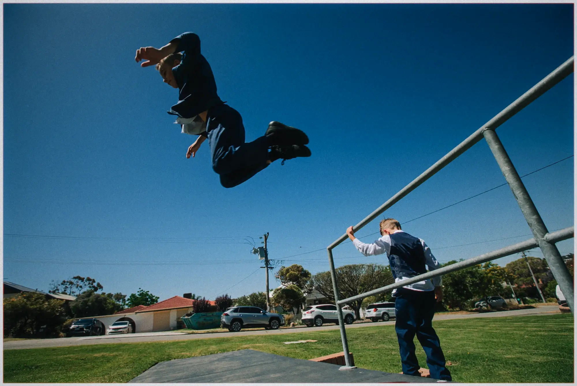 A boy in a navy suit soars off a ramp in a parkour leap under a clear blue sky.