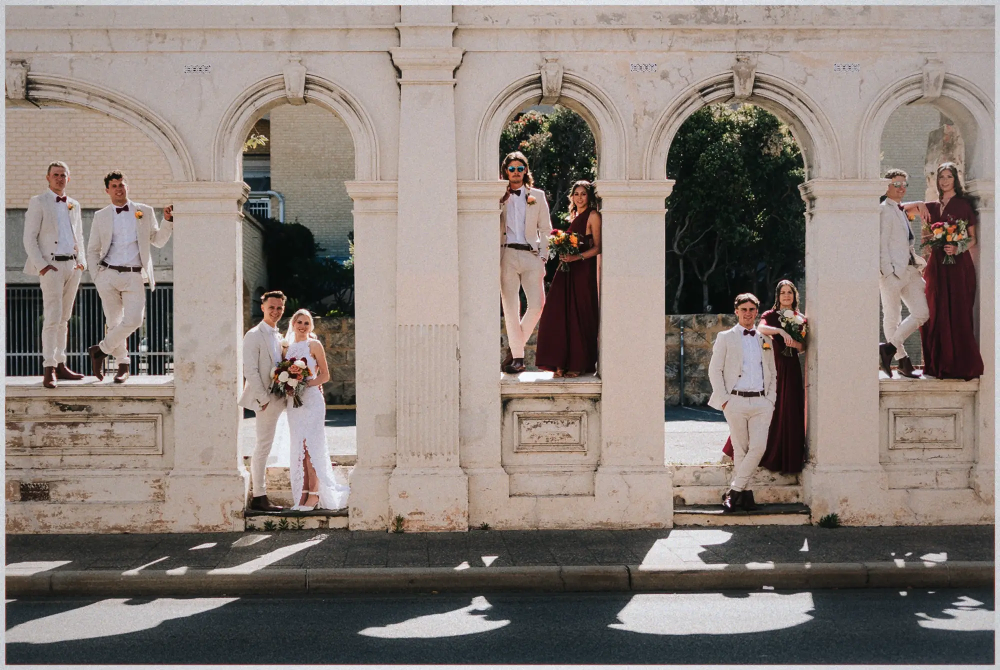Bridal party pairs posing in the sunlit arches of a weathered stone colonnade around Erin and Justin.