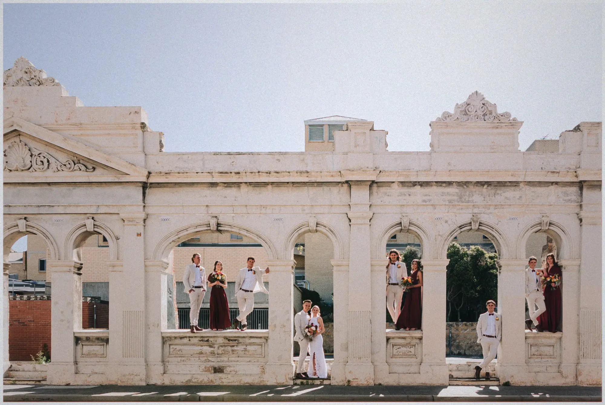 The whole bridal party spread across the weathered white colonnade facade under a pale sky.
