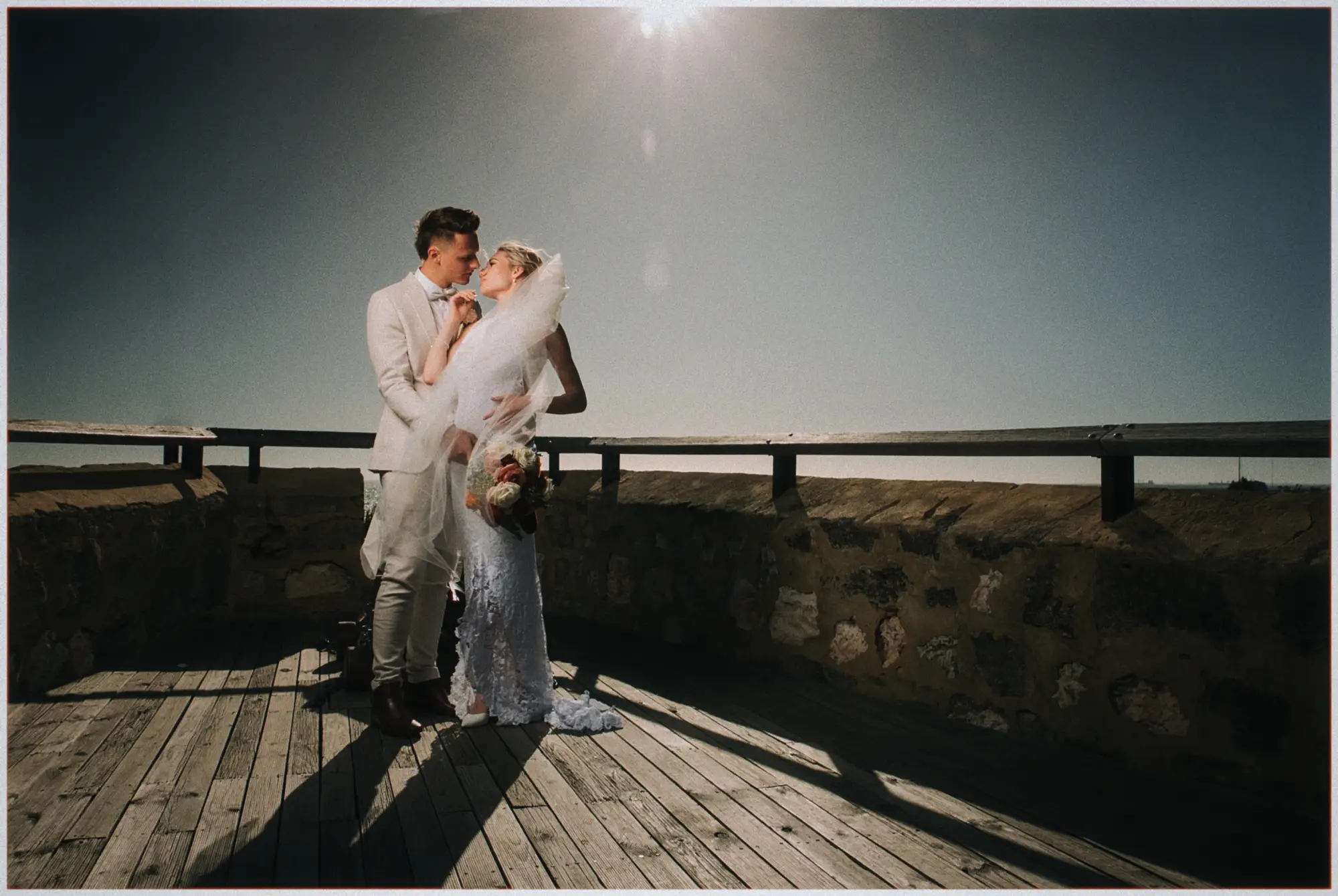 Erin and Justin lean in to kiss on a sunlit timber lookout under a hazy afternoon sun.