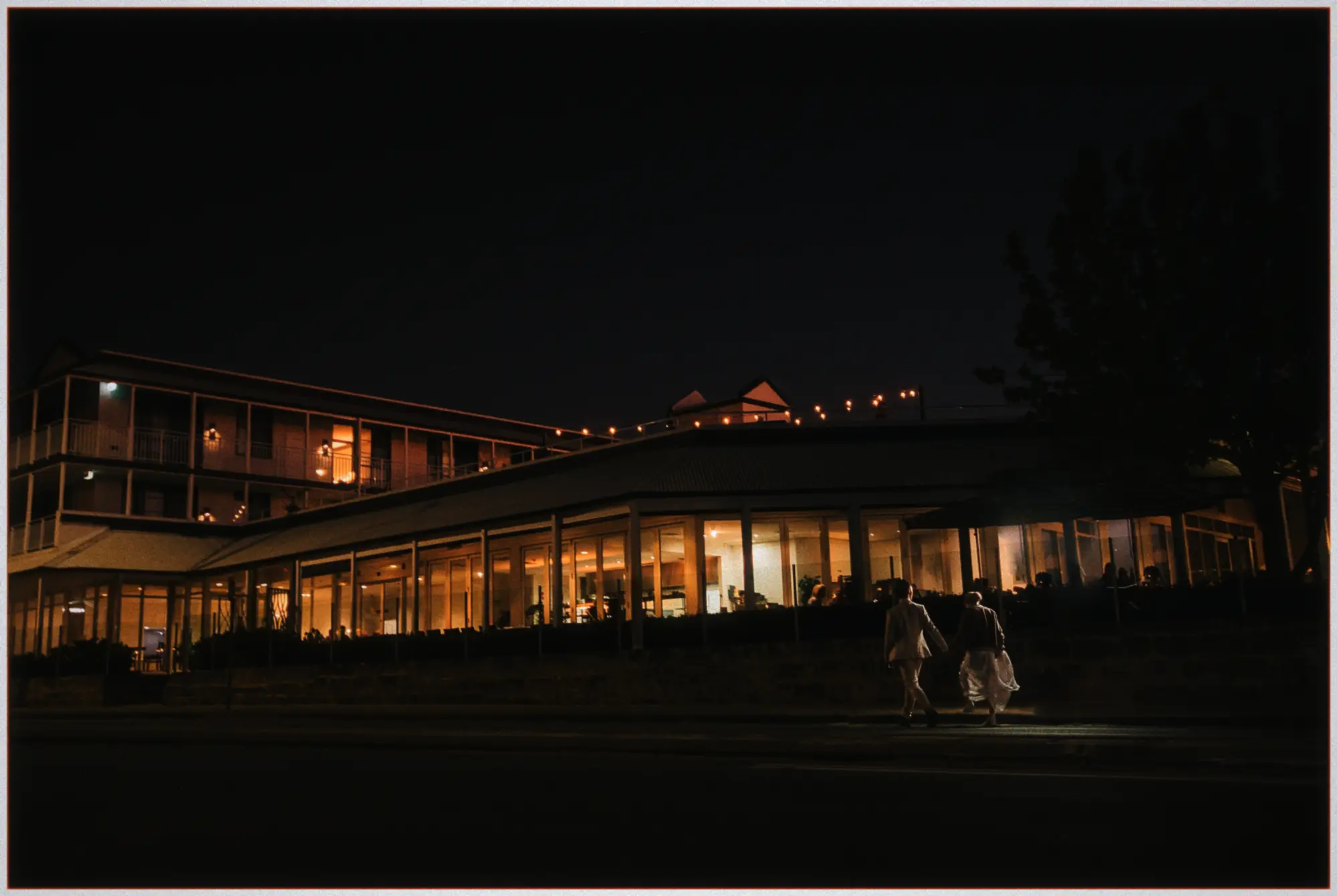 Erin and Justin stroll past the glowing glass-fronted hotel at night beneath festoon lights.
